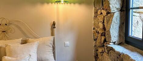 Un dormitorio con cama, almohadas, mesita de noche, pared de piedra y ventana.