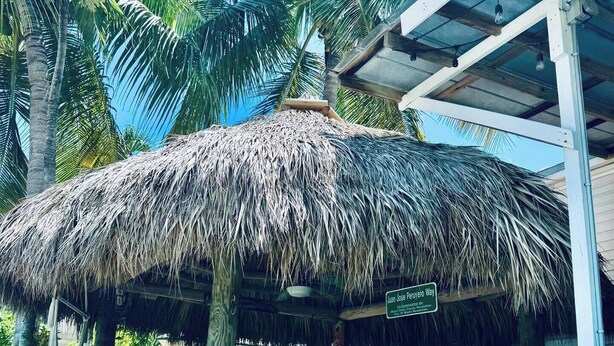 A thatched-roof structure with a sign reading "Juan Jose Guerrero Way" in a tropical setting with palm trees.