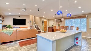 A spacious living room with a large TV, a sofa, and a dining area with a fruit bowl. - 5BR Retreat: Mtn Views, Game Room, Near Broadmoor (Colorado Springs)