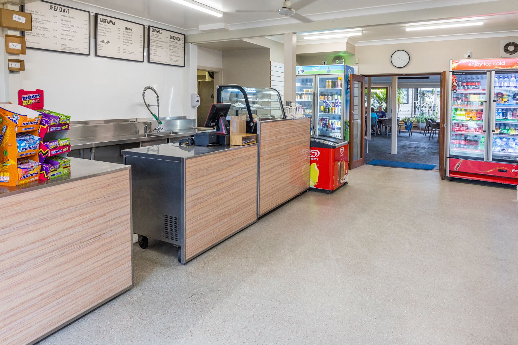 A clean, well-lit food service area with a self-service counter, a beverage dispenser, and a refrigerated display case.