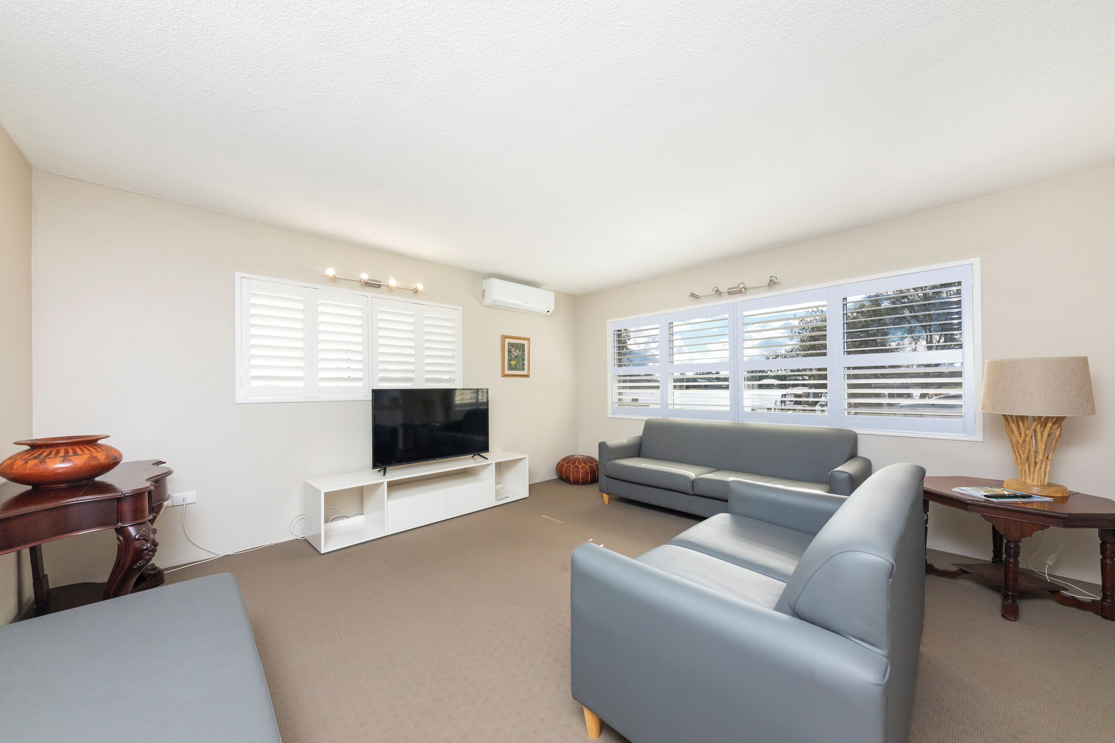 A living room with a grey sofa, a flat-screen TV on a white stand, a wooden side table, and a window with blinds.