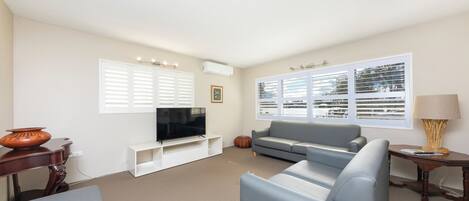 A living room with a grey sofa, a flat-screen TV on a white stand, a wooden side table, and a window with blinds.