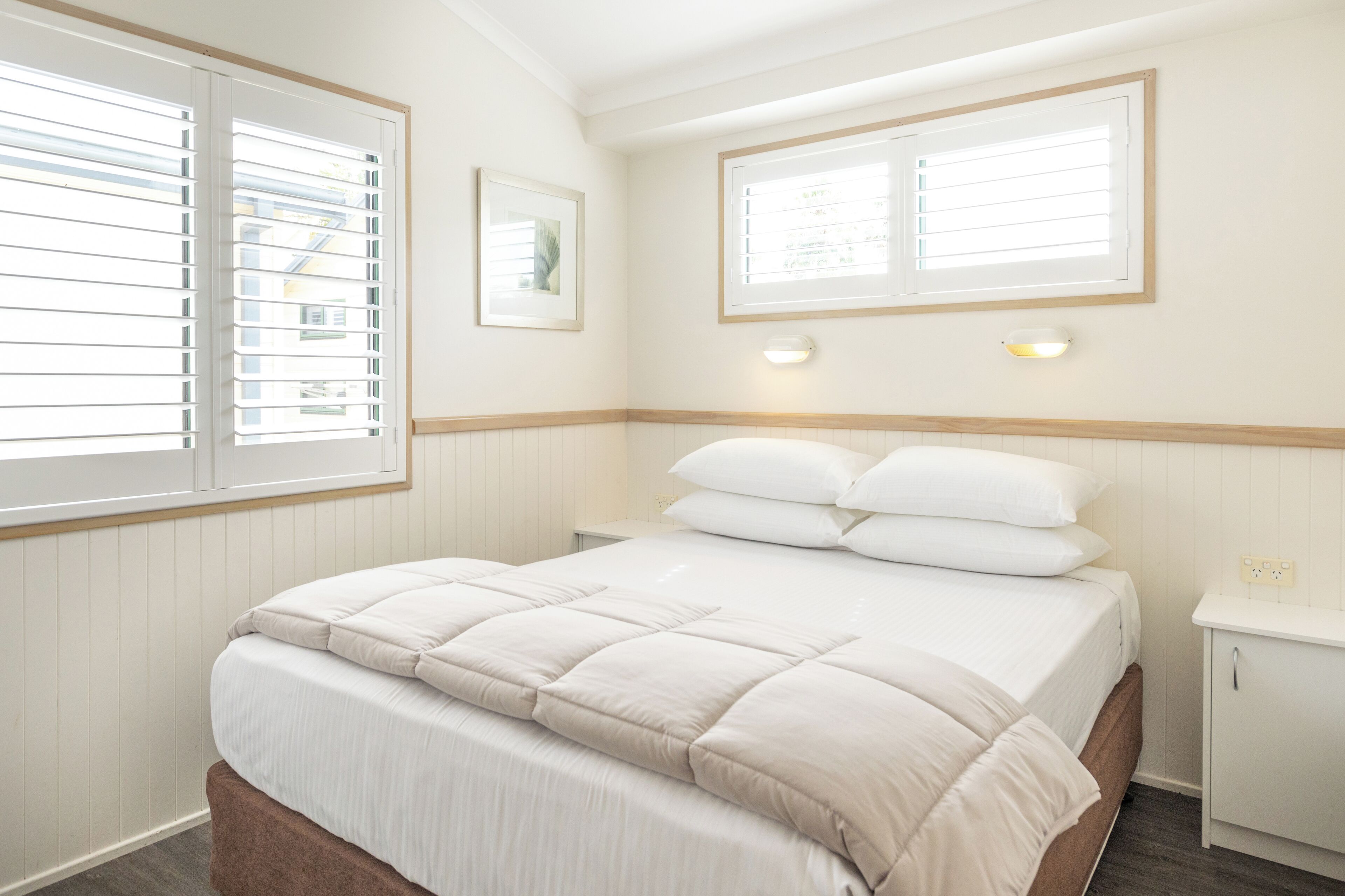 A bedroom with a bed, two bedside tables, a window with blinds, and a wall-mounted light fixture.