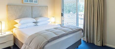 Ein Hotelzimmer mit einem großen Bett, zwei Nachttischen, einem Fenster mit Vorhängen und Blick ins Freie.