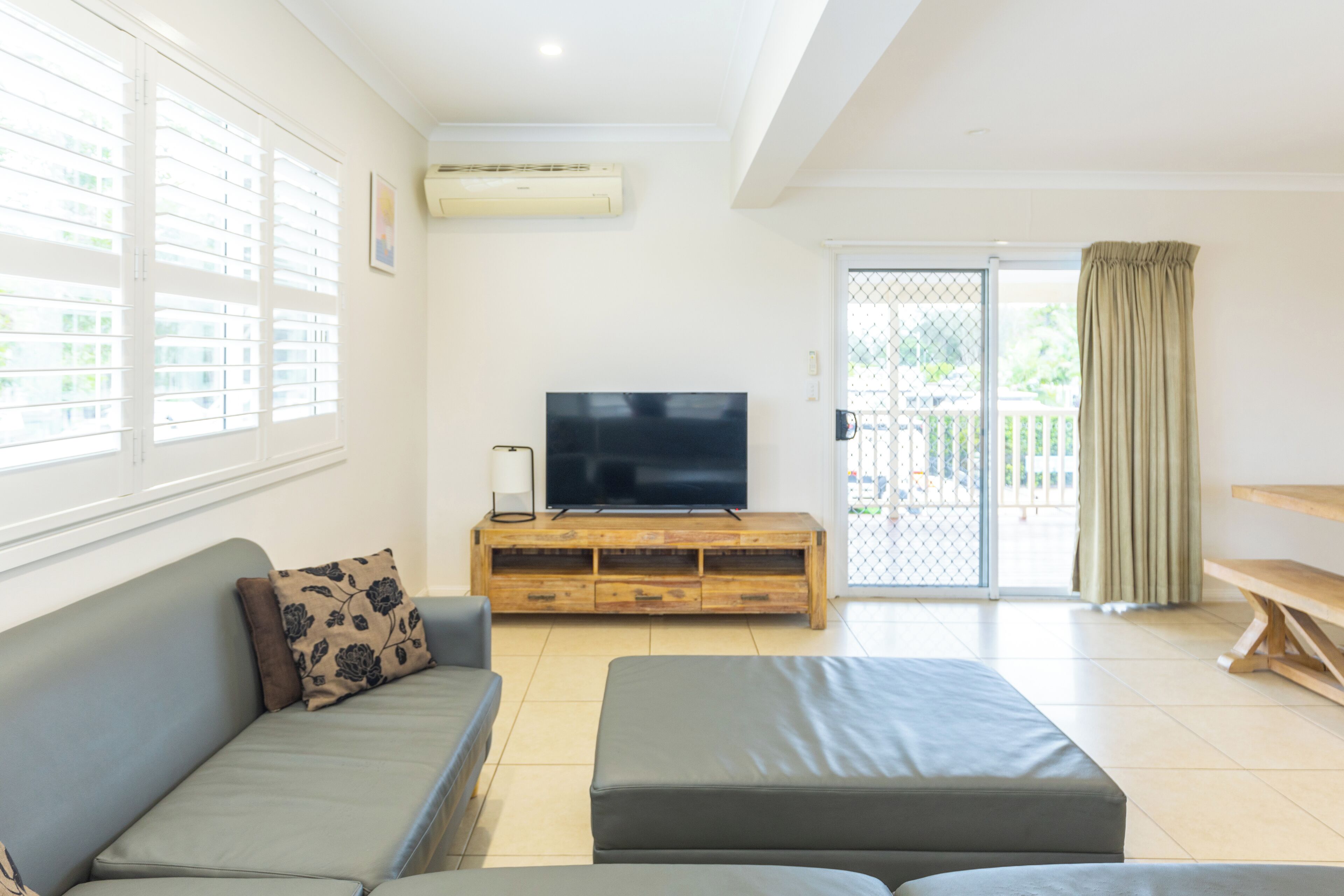 Una sala de estar con un sofá gris, un televisor sobre un mueble de madera y un ventanal corredero que da a un espacio al aire libre.