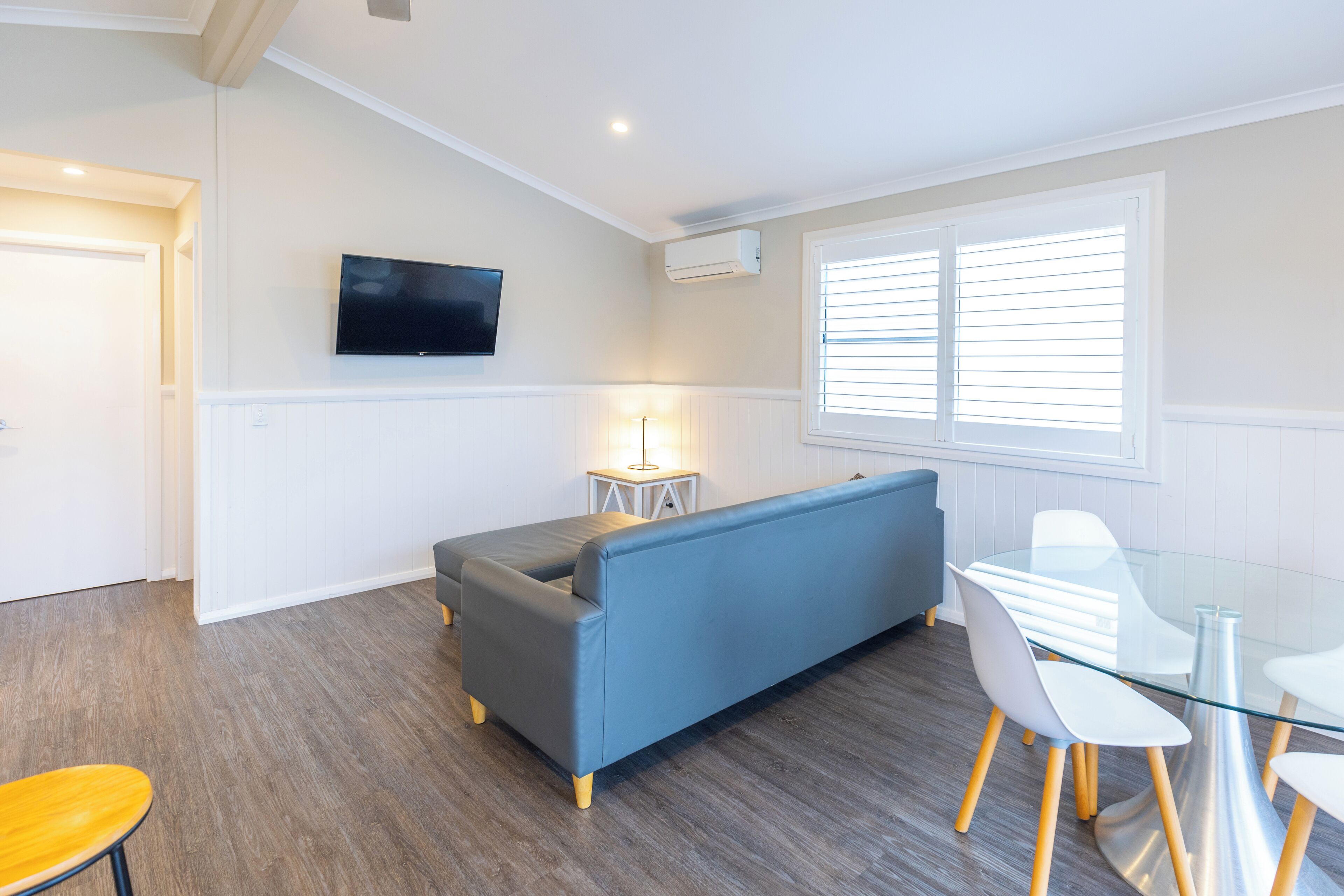 A modern living room with a sofa, a glass dining table, and a TV mounted on the wall.