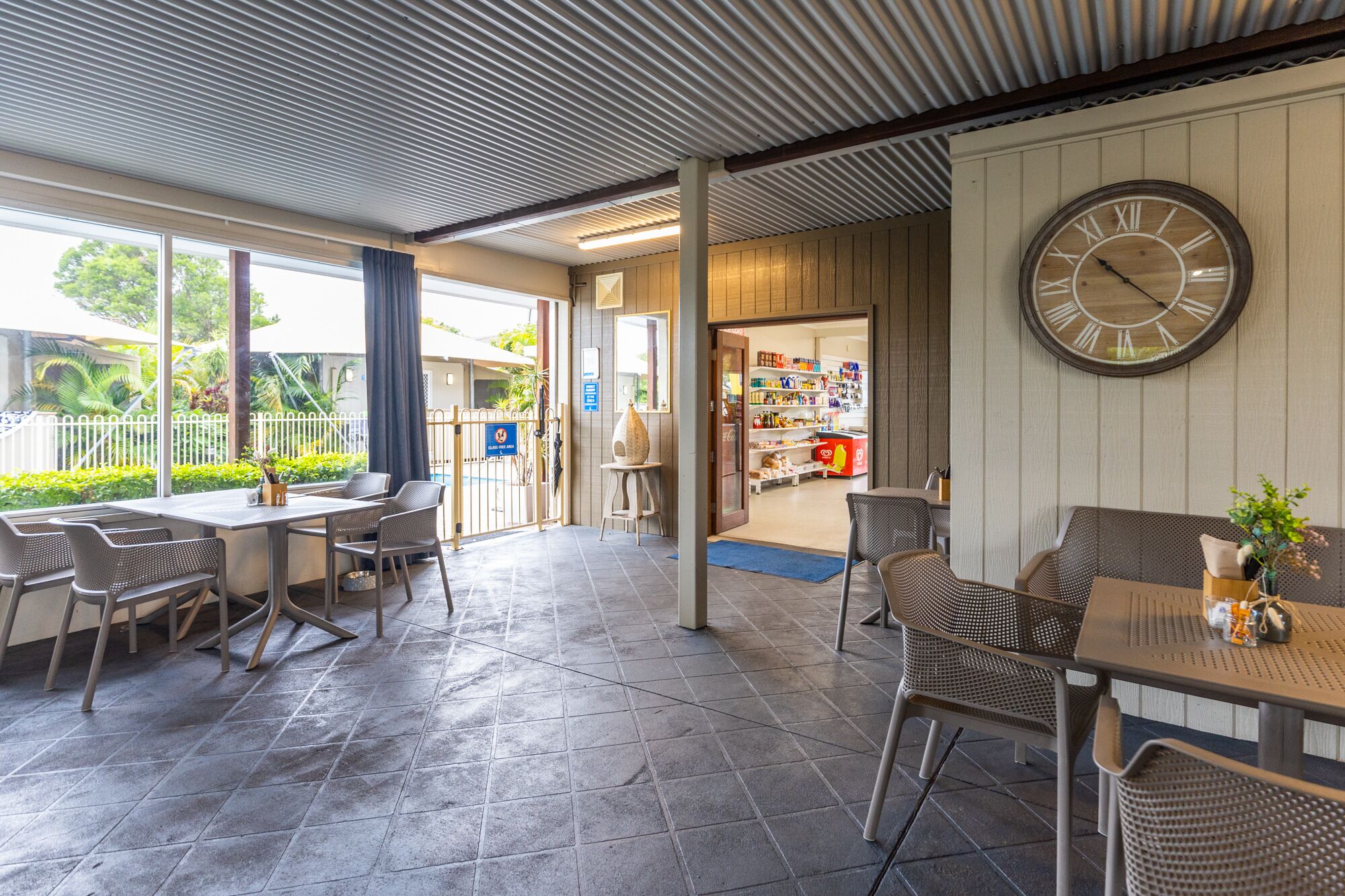 A covered outdoor seating area with tables and chairs, a large wall clock, and a view of a store through an open door.