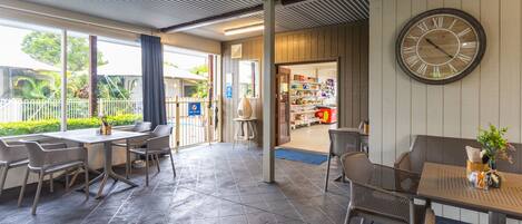 A covered outdoor seating area with tables and chairs, a large wall clock, and a view of a store through an open door.