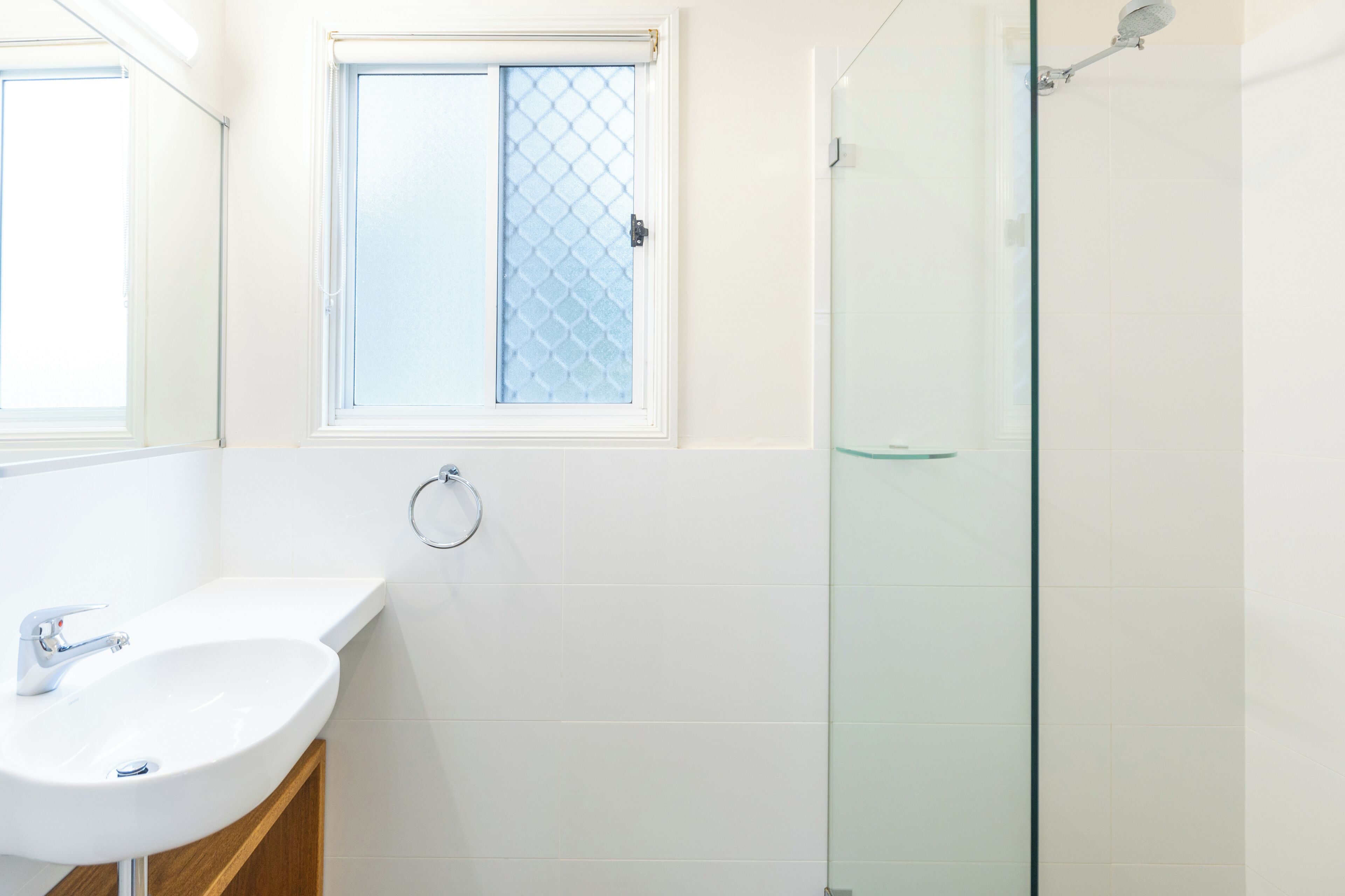 Un baño con lavamanos blanco, cabina de ducha de vidrio y ventana con mosquitera.