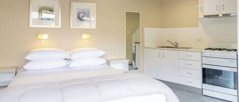 A bedroom with a large bed, a small bedside table, and a nightstand. Adjacent to the bedroom is a kitchenette with a stove, sink, and cabinets.