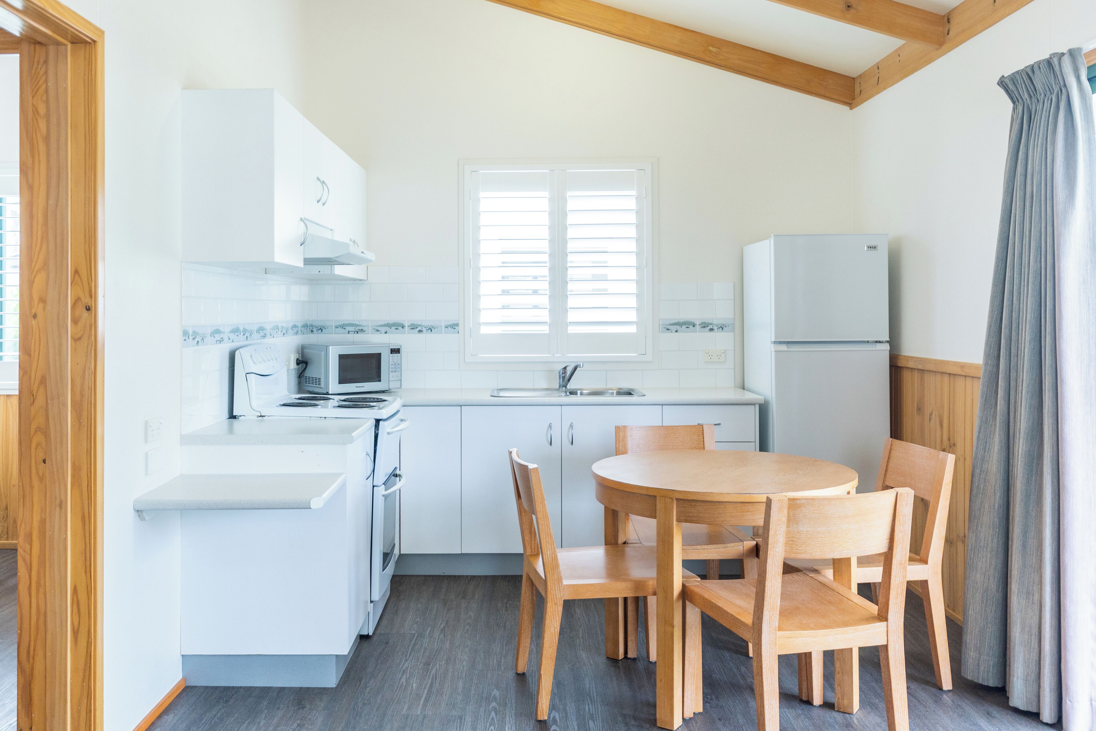Cocina compacta con muebles blancos, microondas, estufa y lavaplatos. Hay una mesa de comedor con cuatro sillas y una ventana con persianas.