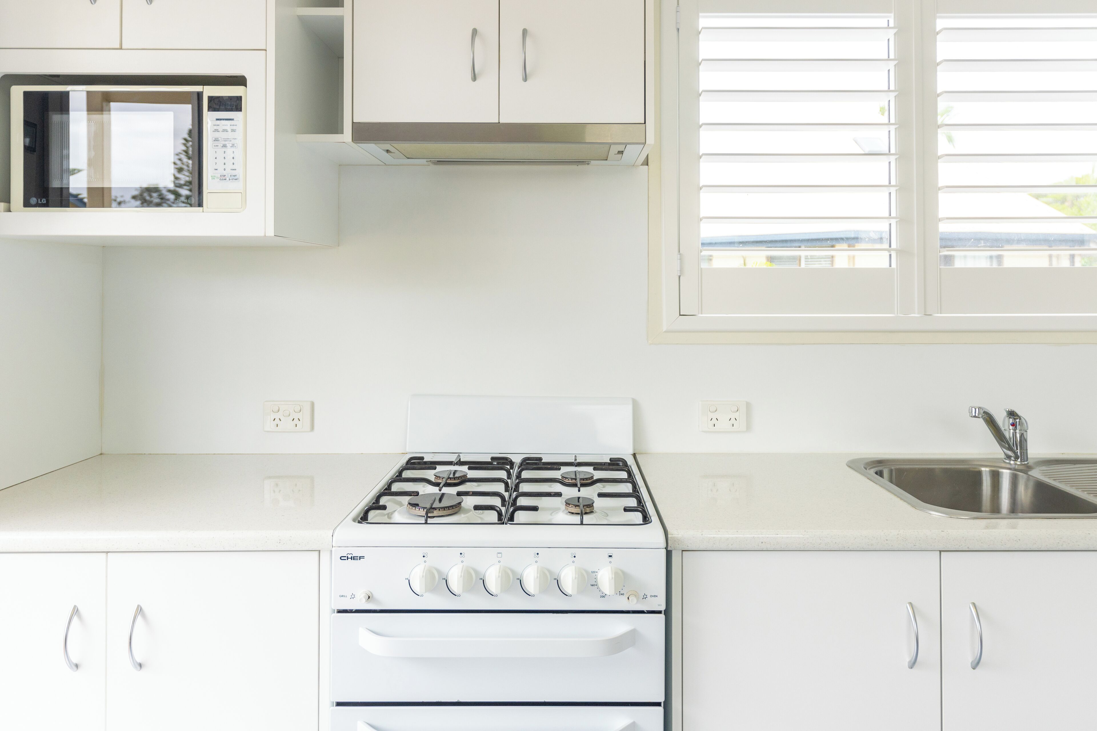 Een moderne keuken met witte kasten, een ingebouwde magnetron, een gasfornuis en een raam met jaloezieën.