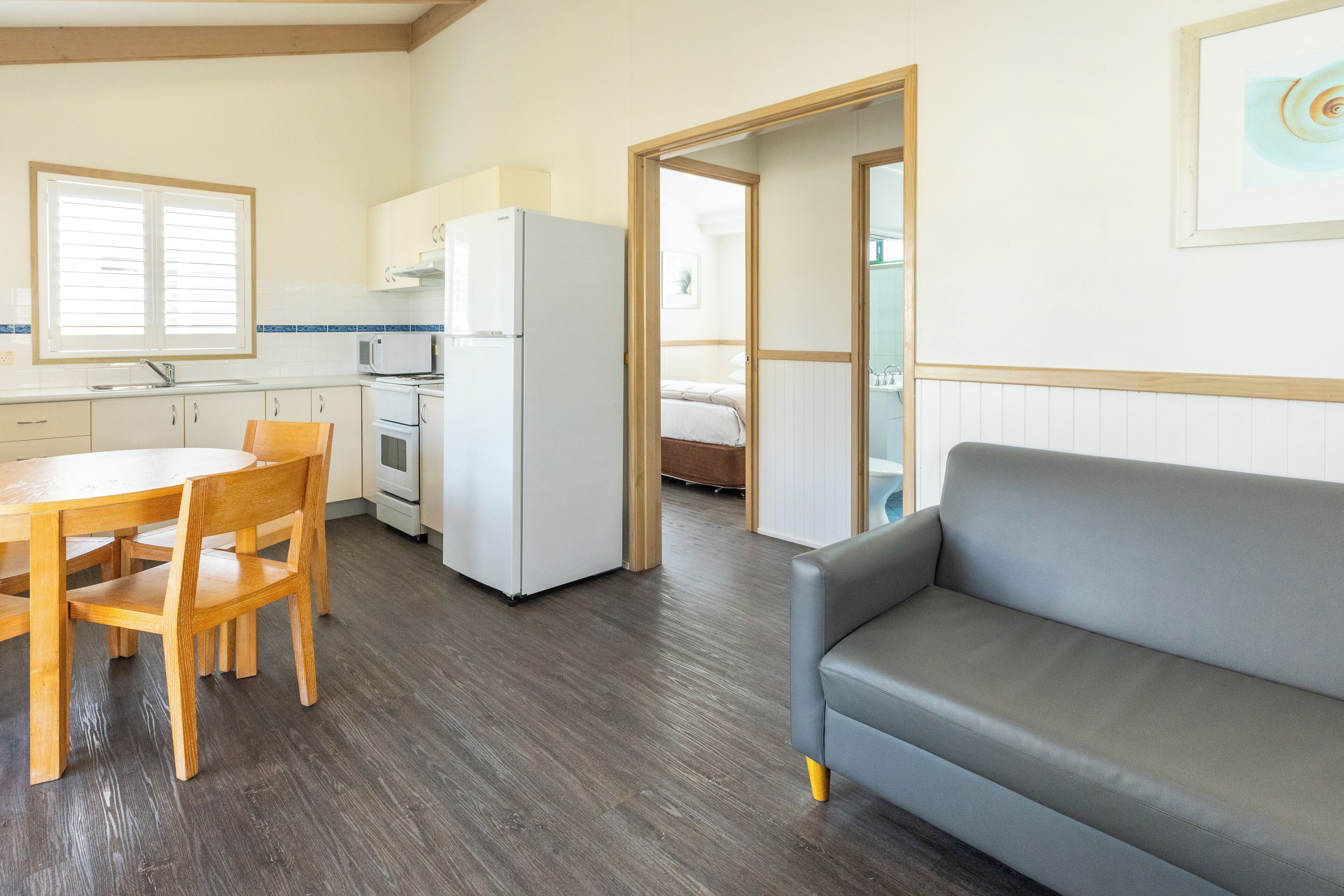 A compact kitchen with a dining table and chairs, a gray sofa, and a visible bed in the adjacent room.