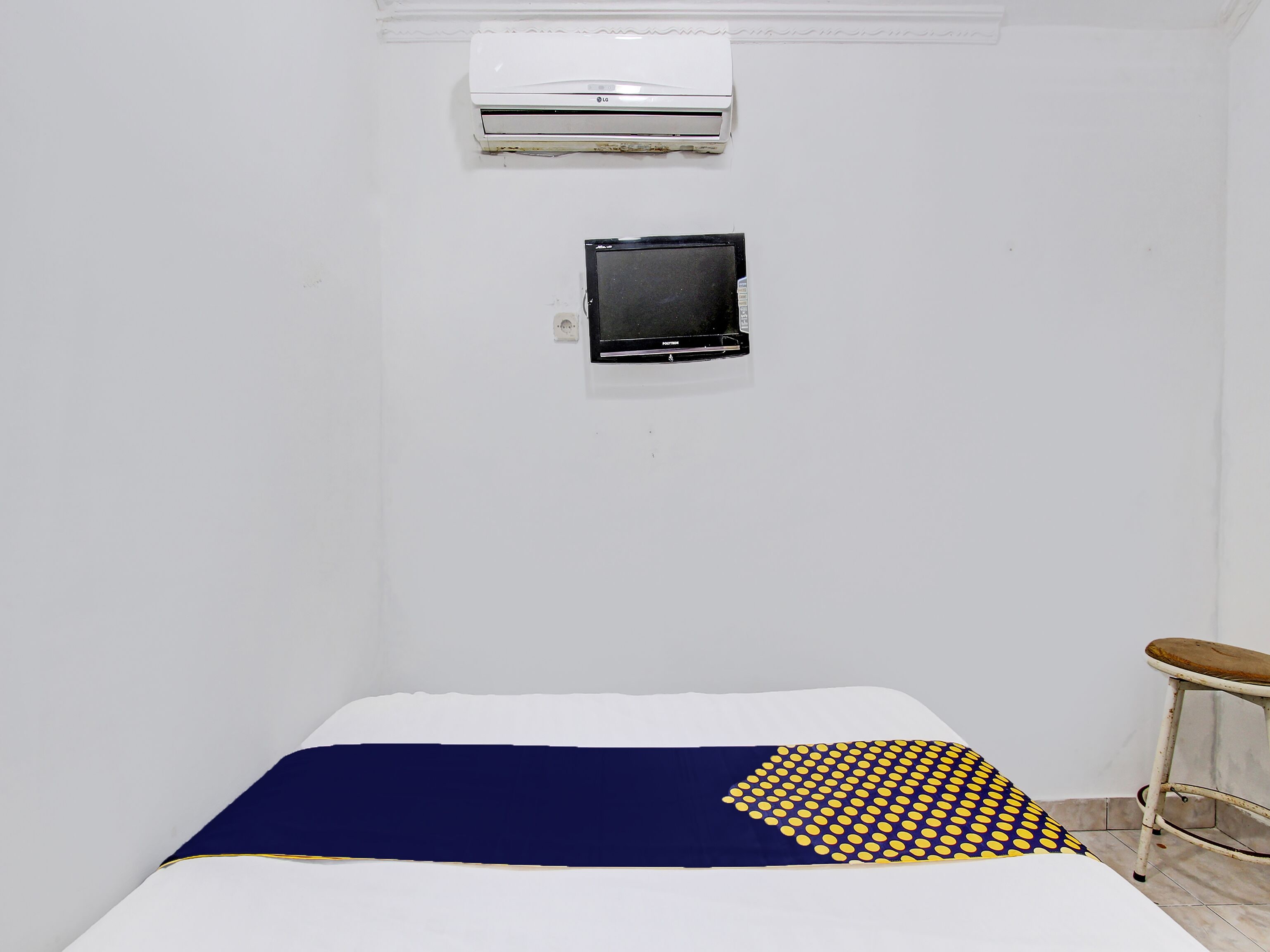 A small, sparsely furnished room with a bed, a small stool, an air conditioner, and a television mounted on the wall.