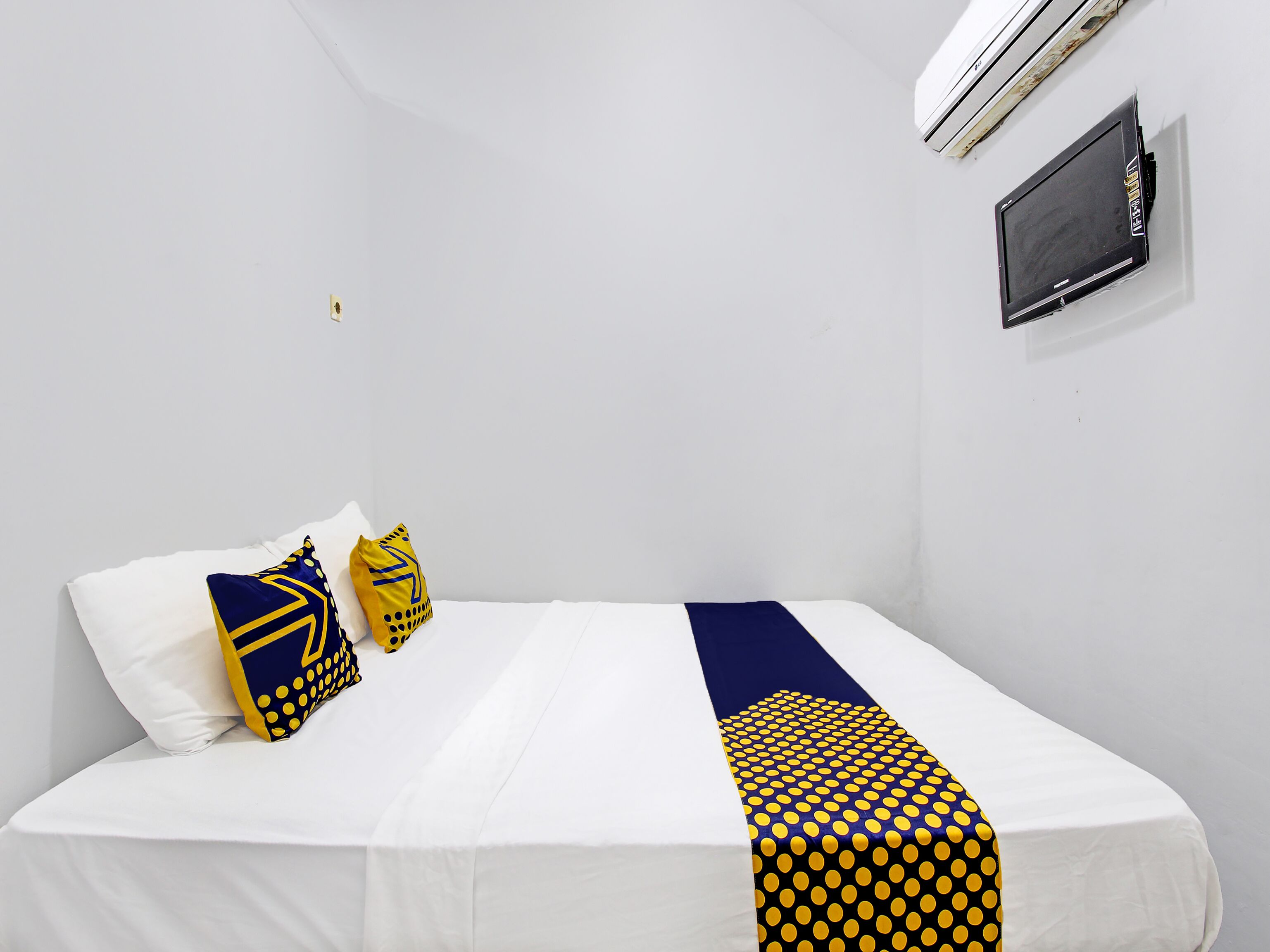 Una cama individual con ropa de cama blanca y un corredor azul y amarillo a rayas. Hay dos almohadas, una blanca y otra con diseños azules y amarillos. Un equipo de aire acondicionado negro está montado en la pared sobre la cama.