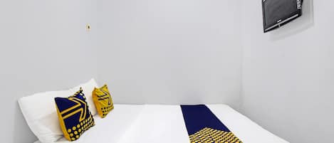 Una cama individual con ropa de cama blanca y un corredor azul y amarillo a rayas. Hay dos almohadas, una blanca y otra con diseños azules y amarillos. Un equipo de aire acondicionado negro está montado en la pared sobre la cama.
