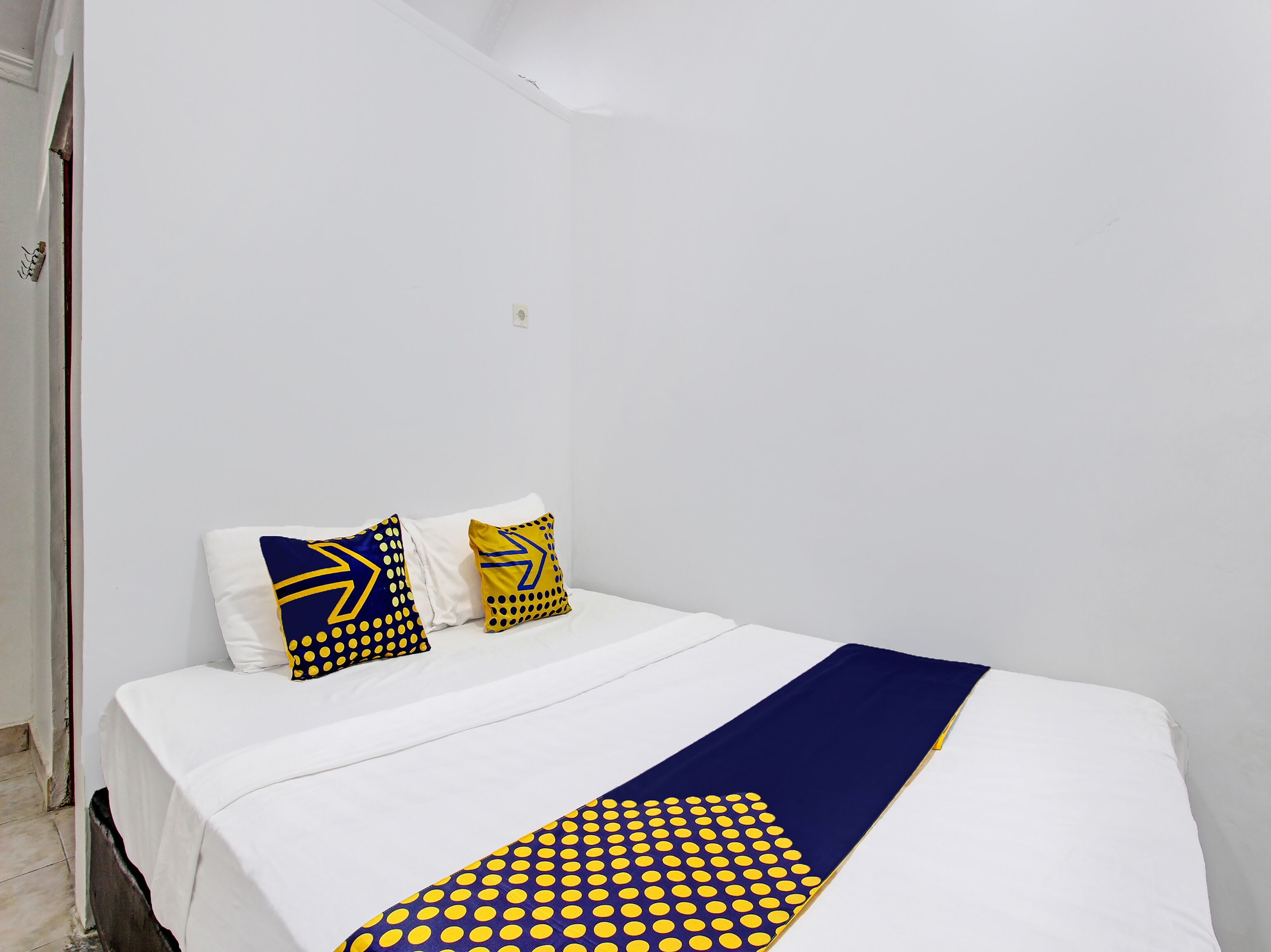 A single-bed room with a white bedspread, two pillows with yellow and blue patterns, and a blue and yellow blanket.