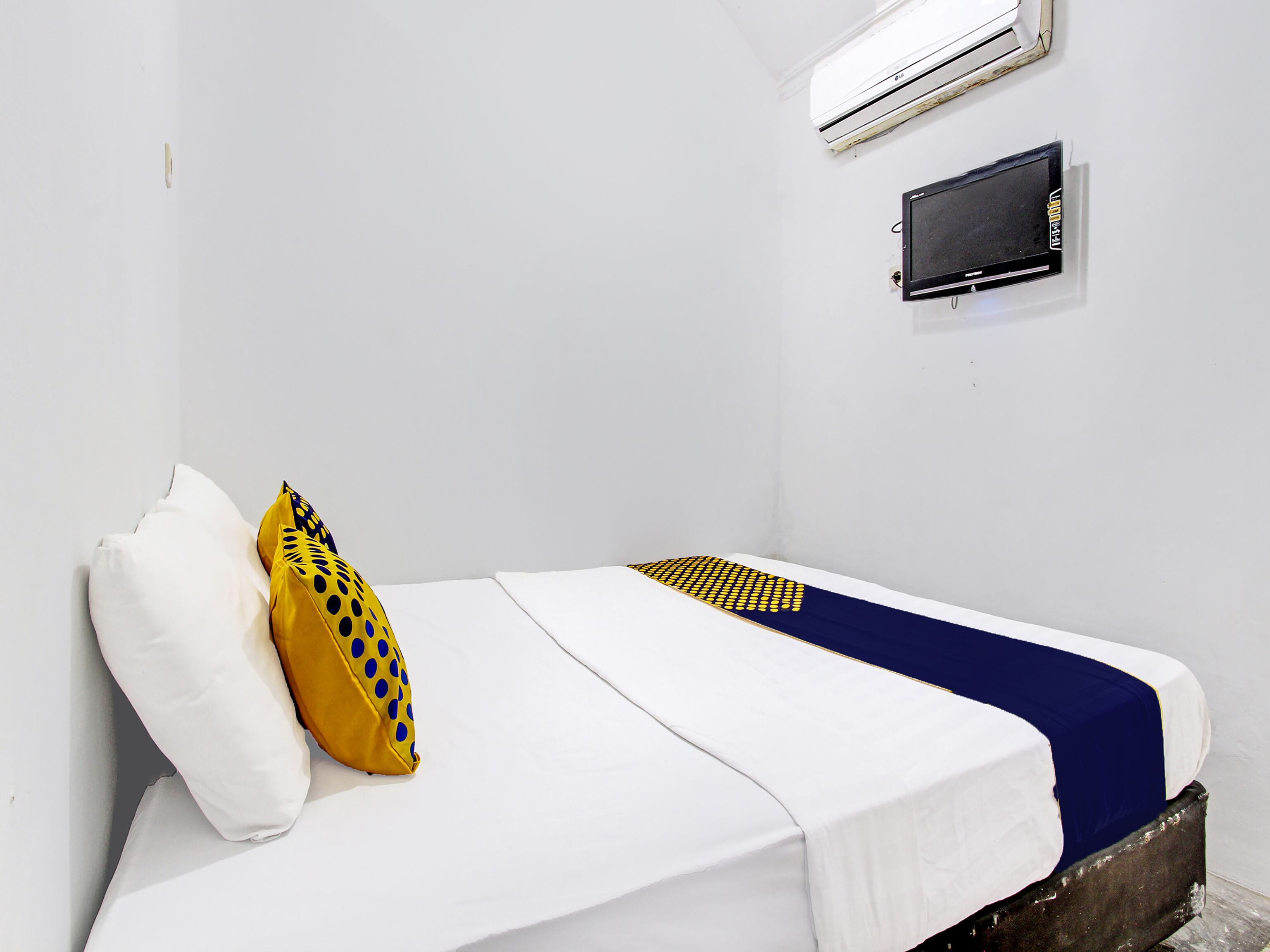 Un lit simple avec une literie blanche et bleue, un oreiller jaune à pois bleus et un petit écran noir accroché au mur.