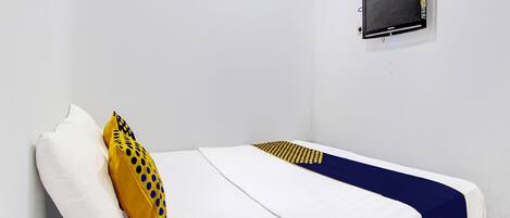 Una cama individual con ropa de cama blanca y azul, un almohón amarillo con lunares azules y una pequeña pantalla negra en la pared.