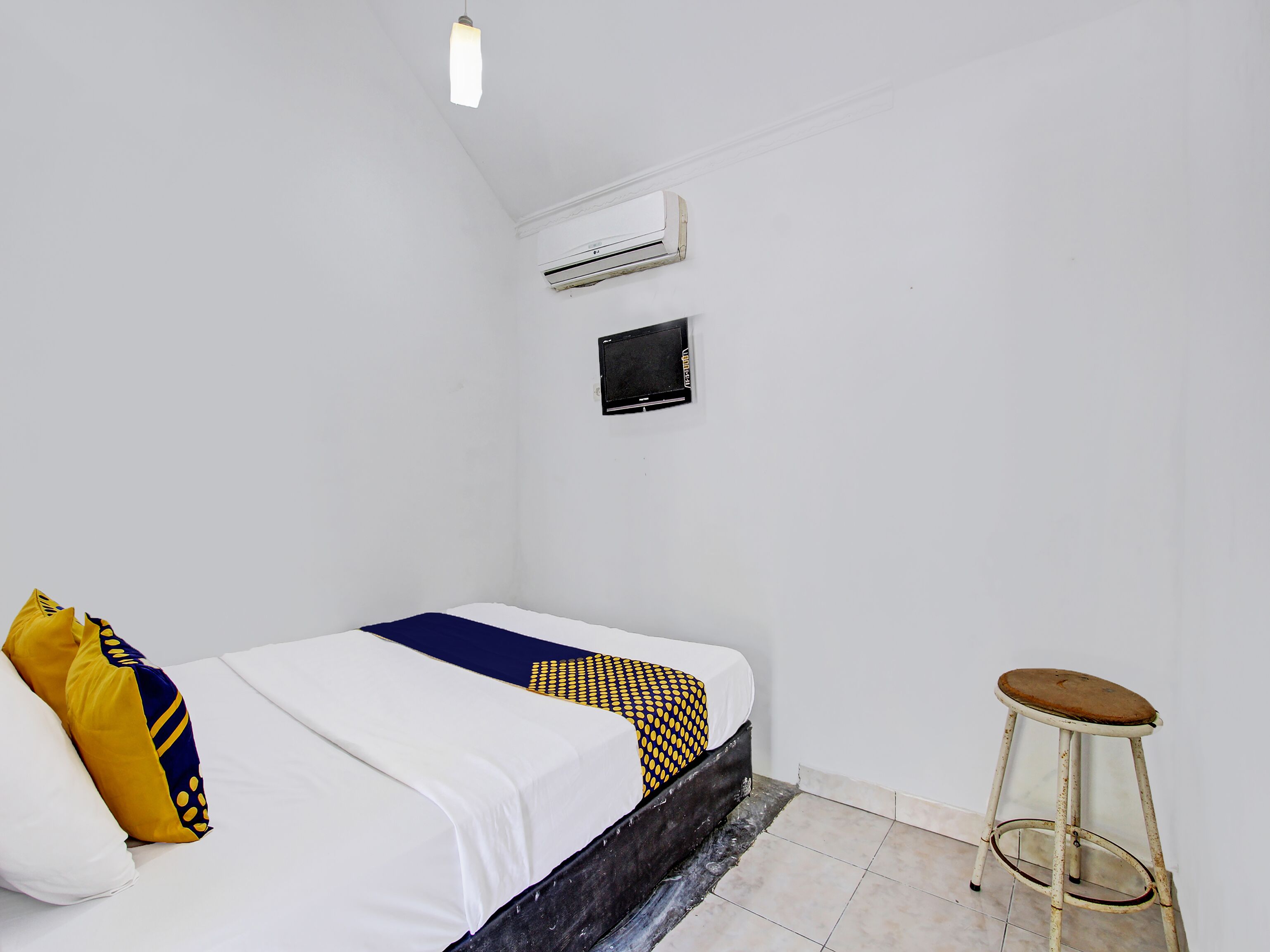 A small, clean room with a bed, a single stool, an air conditioning unit, and a television.
