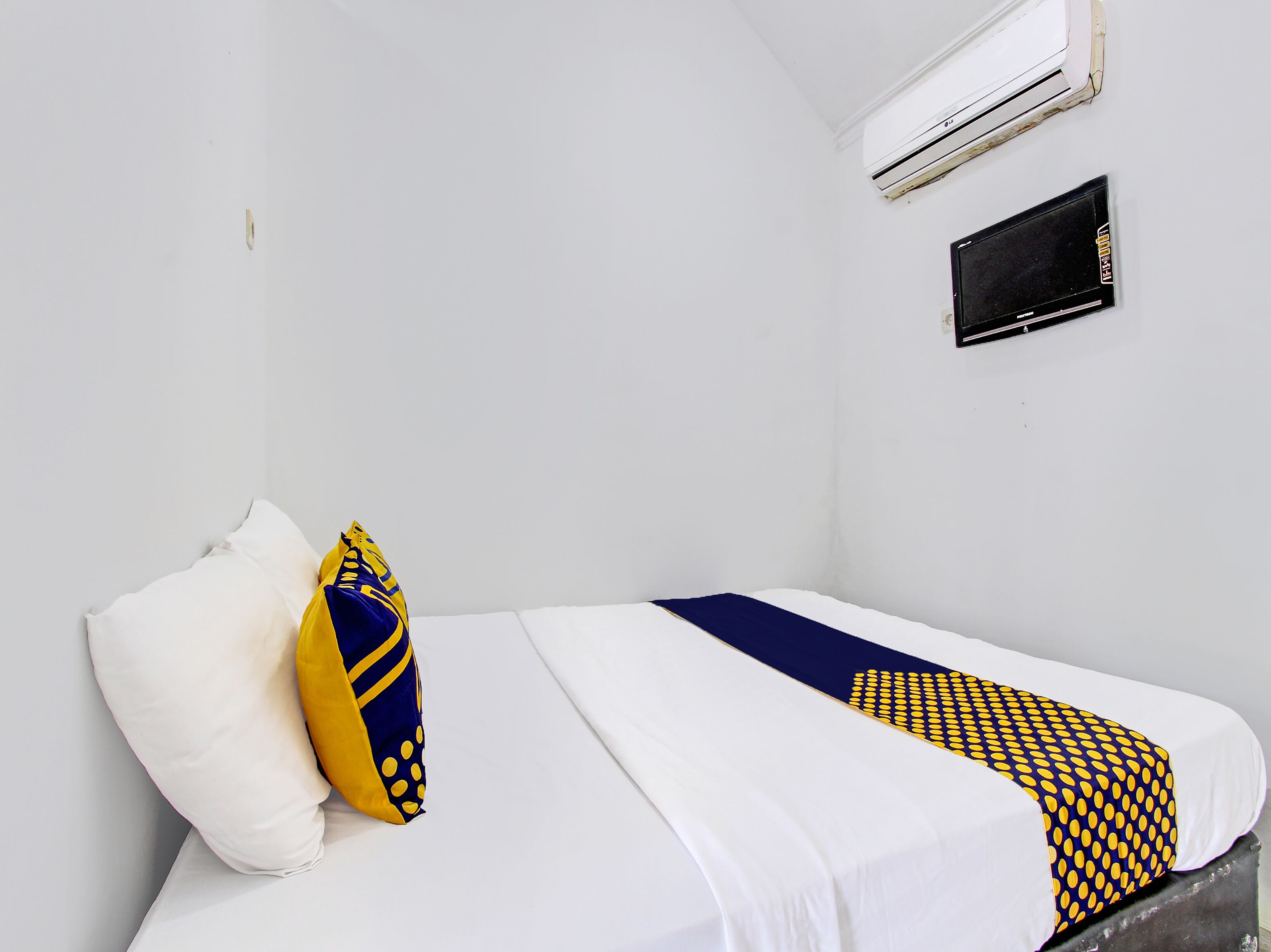 A single bed with white bedding and a blue and yellow patterned blanket. There is a white pillow and a yellow and blue pillow on the bed. An air conditioning unit is mounted on the wall above the bed.