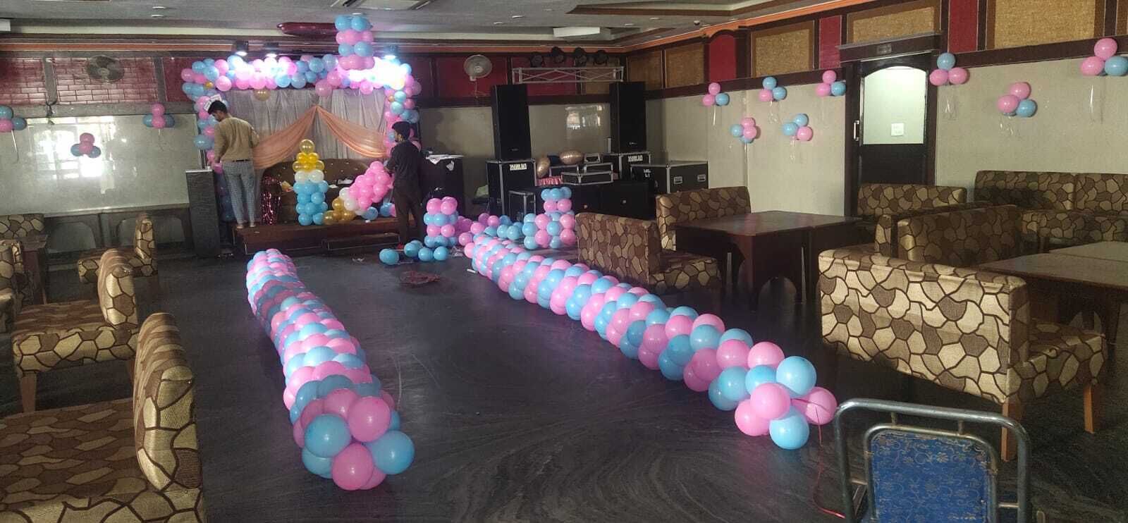 Un salón de banquetes con una larga hilera de globos rosa y azules, sillas con diseño de piedra y un escenario con fondo decorativo.