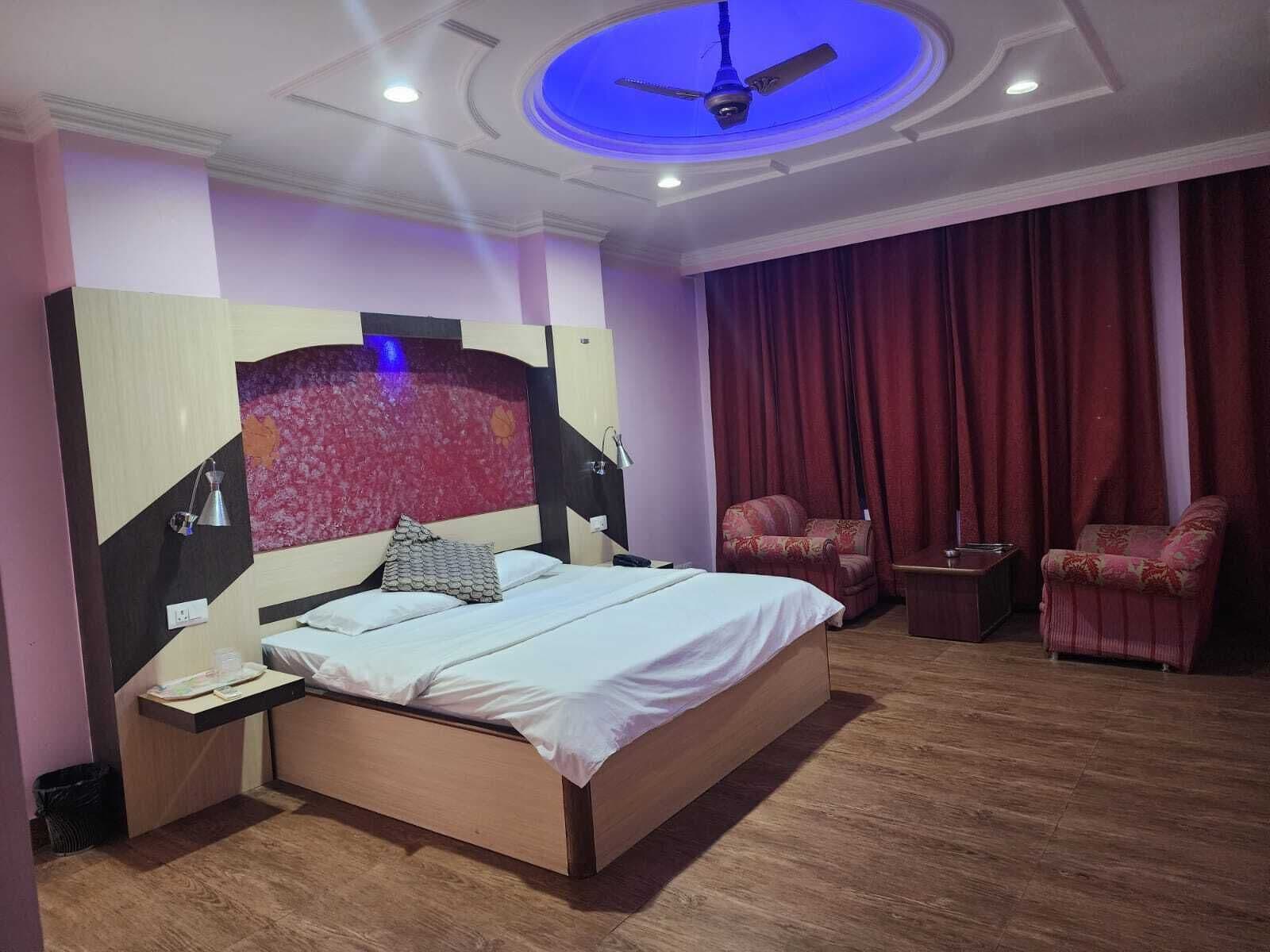 A bedroom with a bed, a desk, two chairs, a ceiling fan, and a large window with curtains.