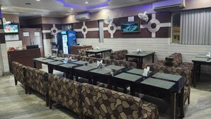A restaurant with a stone-patterned bench seating, tables with green placemats, and a TV screen on the wall. - Hotel Meridian Palace (Jammu)