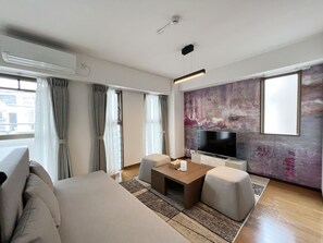 A modern living room with a sofa, coffee table, and TV. There is a large window with curtains, an air conditioning unit, and a wall-mounted light fixture. - Dio Higashi Shinsaibashi (Osaka)