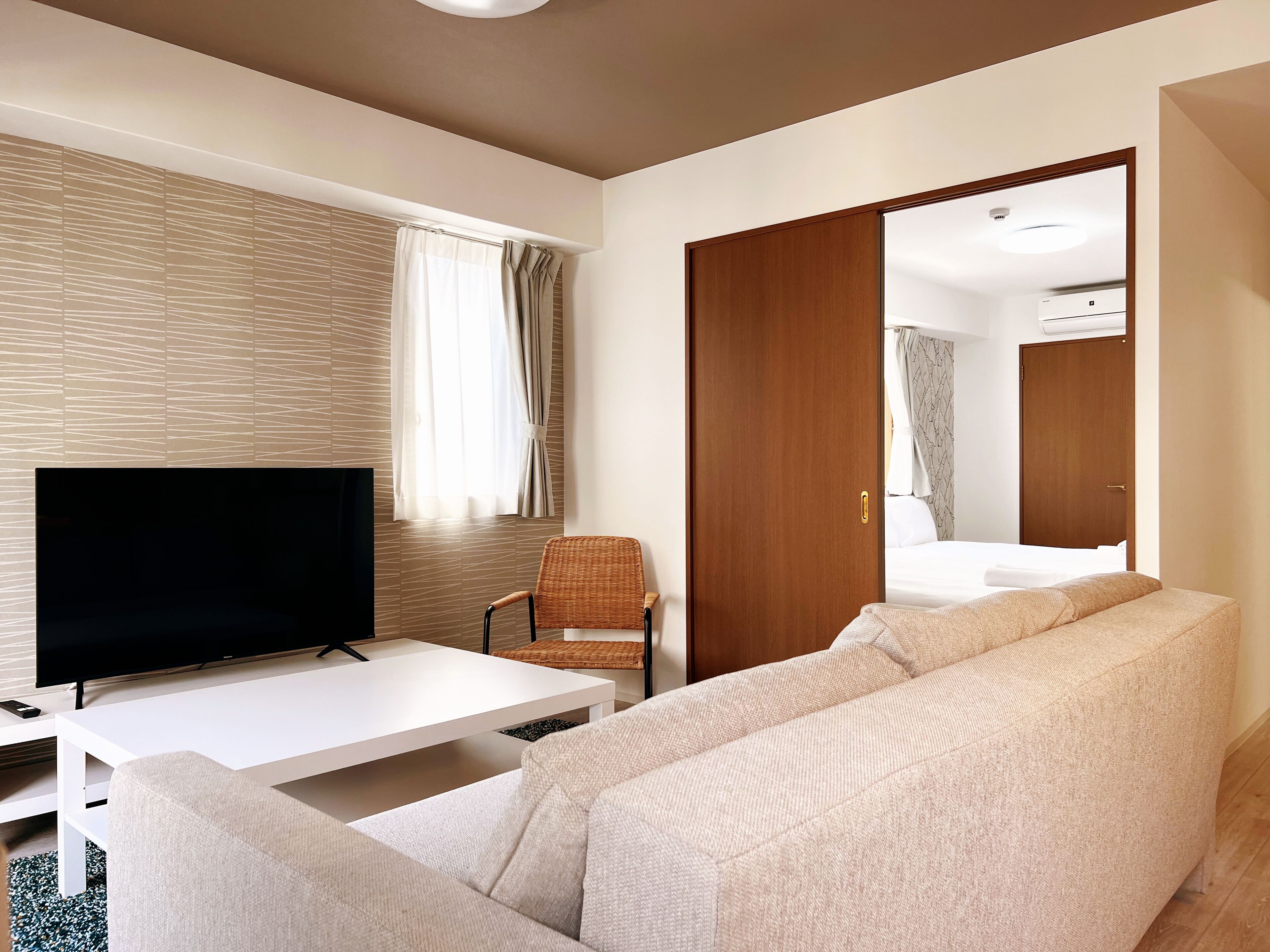 Een moderne hotelkamer met een flatscreen televisie, een witte bank, een rieten fauteuil en een houten deur die leidt naar een badkamer.