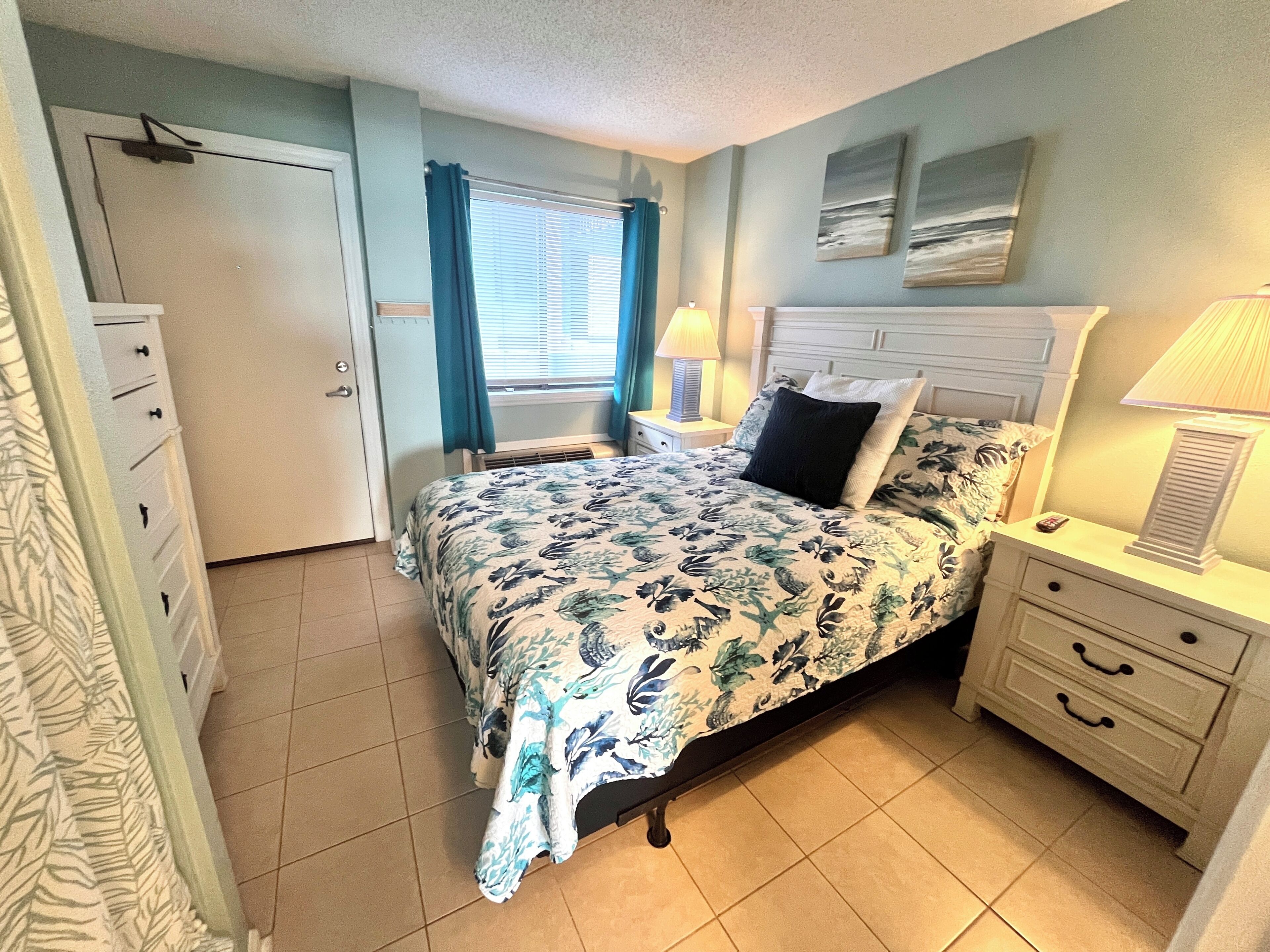 A bedroom with a bed, bedside tables, a nightstand, a window with curtains, and a door.
