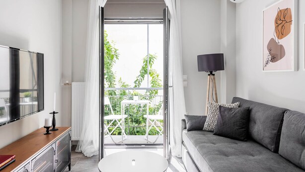 Un salón moderno con un sofá gris, una mesa redonda blanca y un televisor a la izquierda. Se ve un balcón con plantas a través de la ventana.