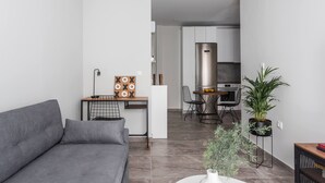 Una sala de estar moderna con un sofá gris, una pequeña mesa de madera con un tablero de ajedrez, y un comedor con un refrigerador de acero inoxidable y sillas.