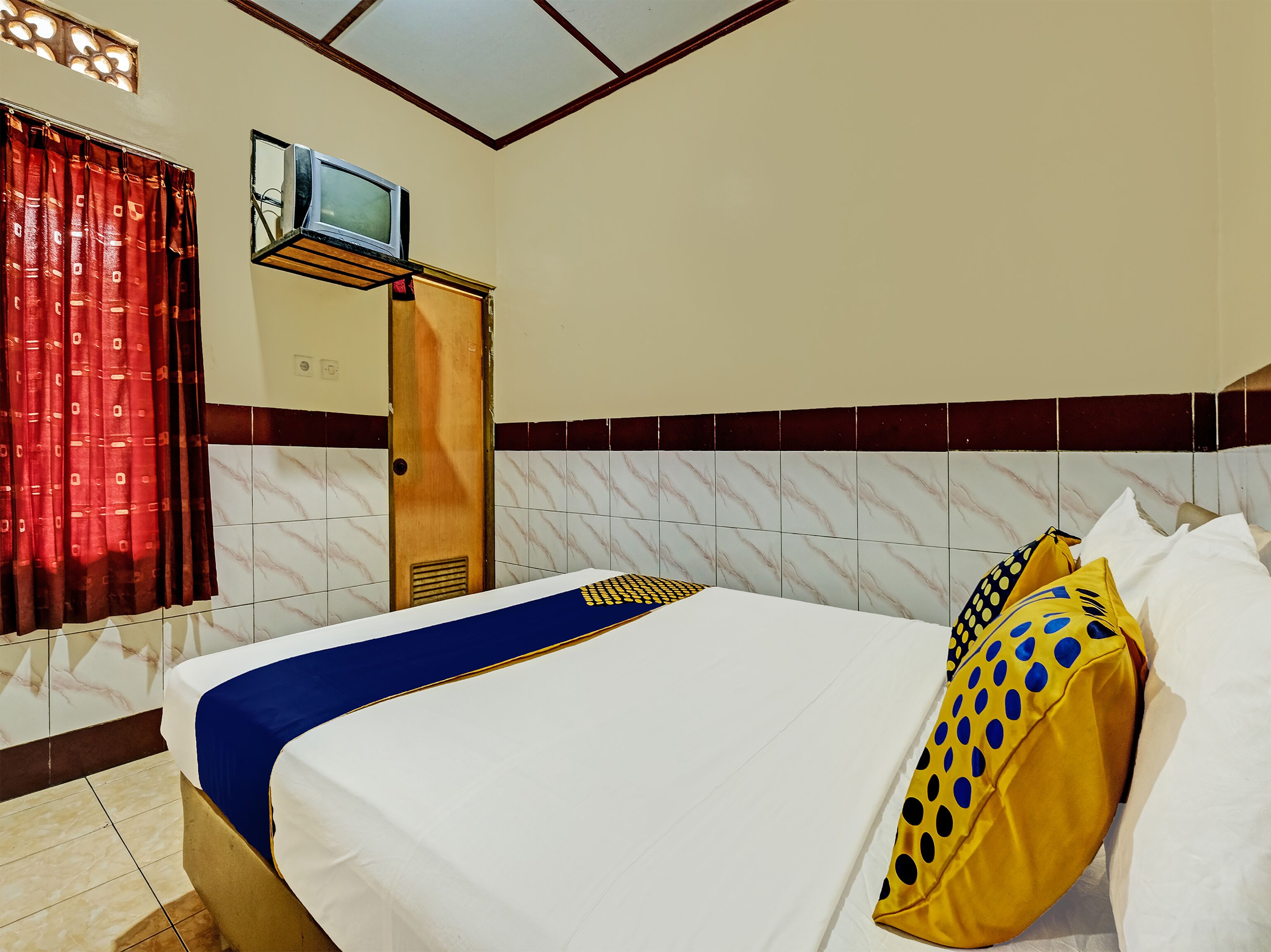 Ein Hotelzimmer mit einem Bett, einem an der Wand befestigten Fernseher und einem Fenster mit roten Vorhängen.