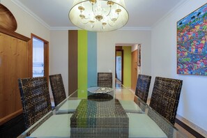 A dining area with a glass table, wicker chairs, a chandelier, and a colorful painting on the wall.