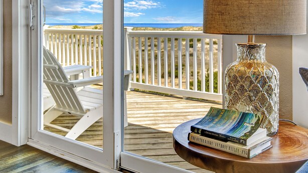 Una habitación con vista a la playa, una silla blanca, una lámpara y libros sobre una mesa.