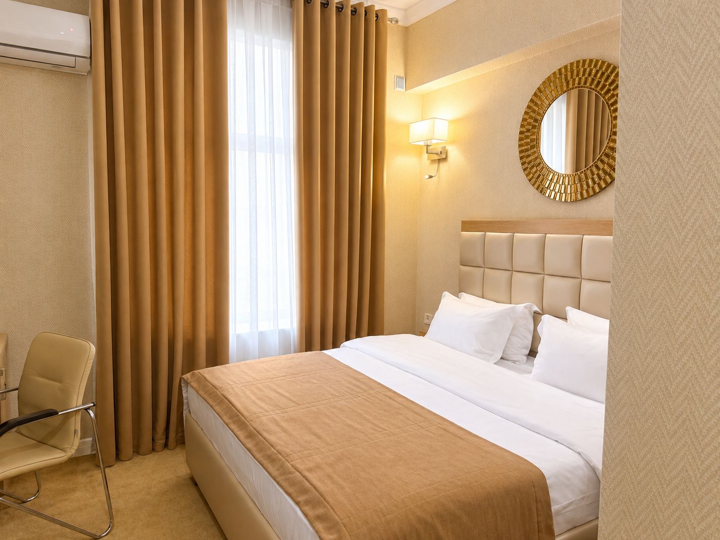 A hotel room with a bed, a chair, a wall-mounted mirror, and a window with curtains.