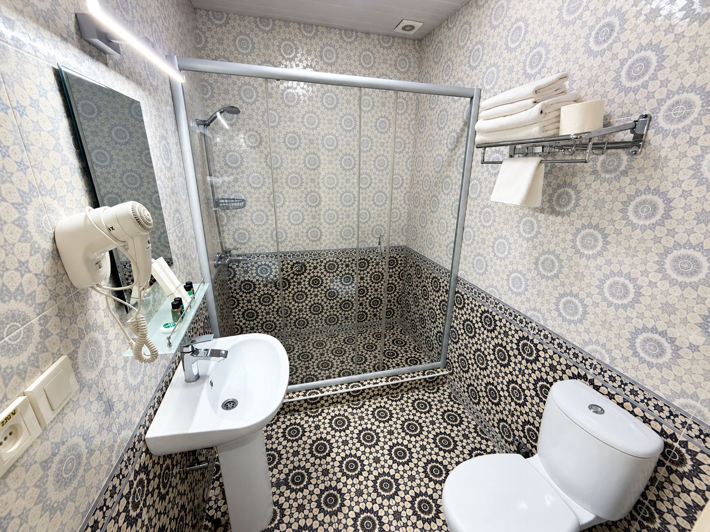 A bathroom with a shower, sink, and toilet, featuring patterned tiles and a towel rack.