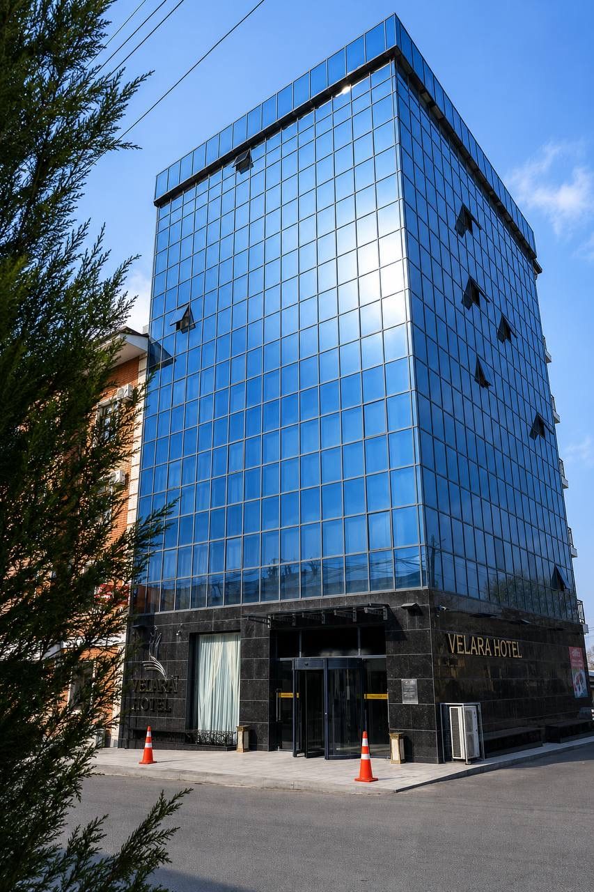 A modern hotel building with a reflective glass facade, a prominent entrance with a black awning, and the name 'VELARA HOTEL' displayed on the side.