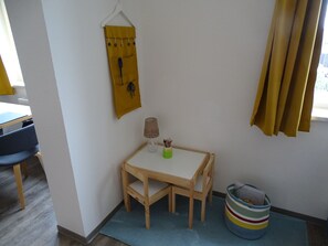 Une petite pièce avec une table et des chaises en bois, une lampe, un sac jaune suspendu et un panier à rayures posé sur le sol.
