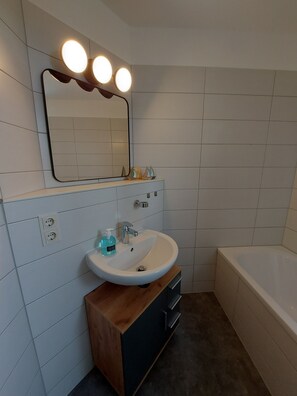 Une salle de bain avec un lavabo blanc, un miroir doté de trois lampes et une baignoire.