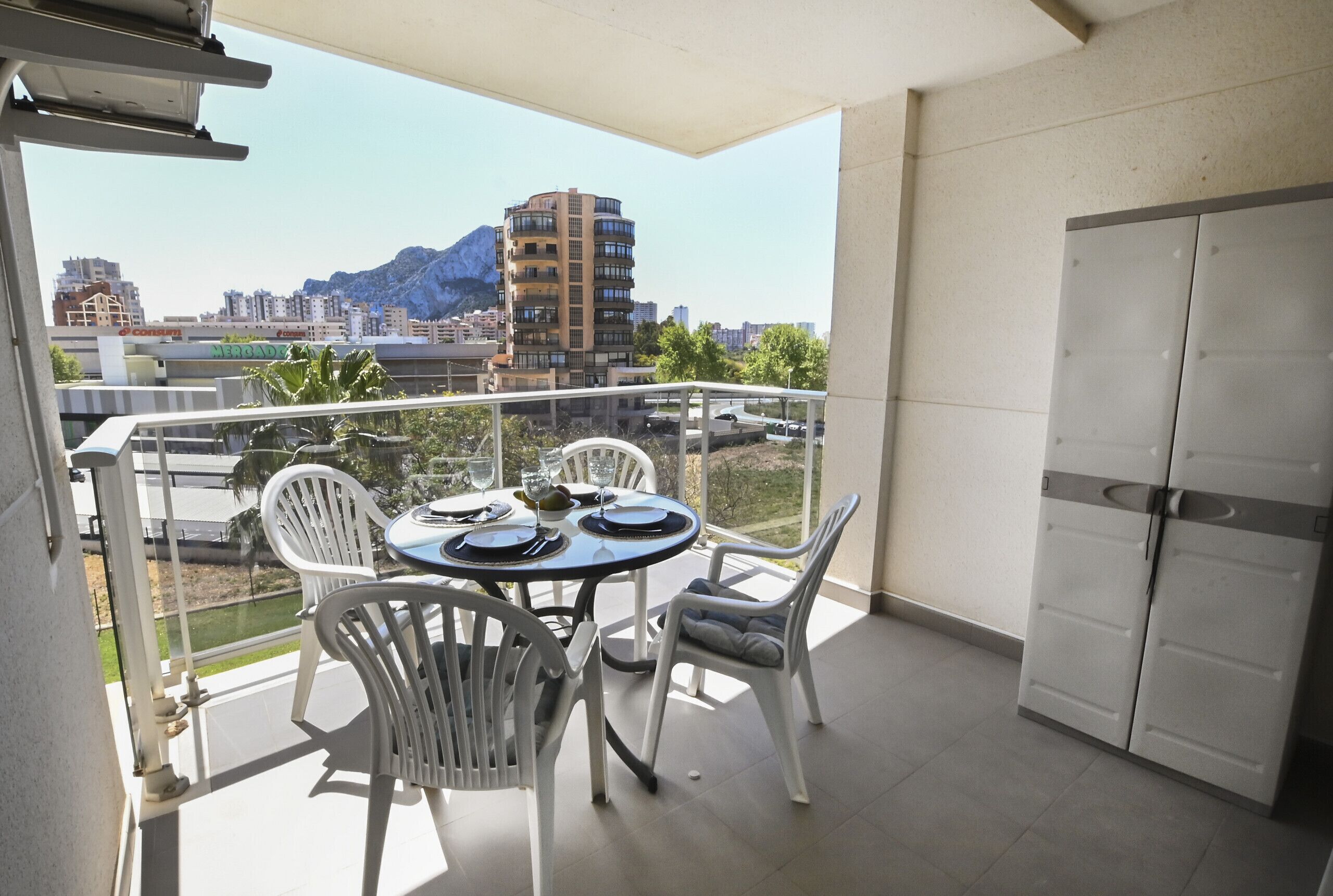 A balcony with a table and chairs, a door, and a view of a cityscape with buildings and mountains.
