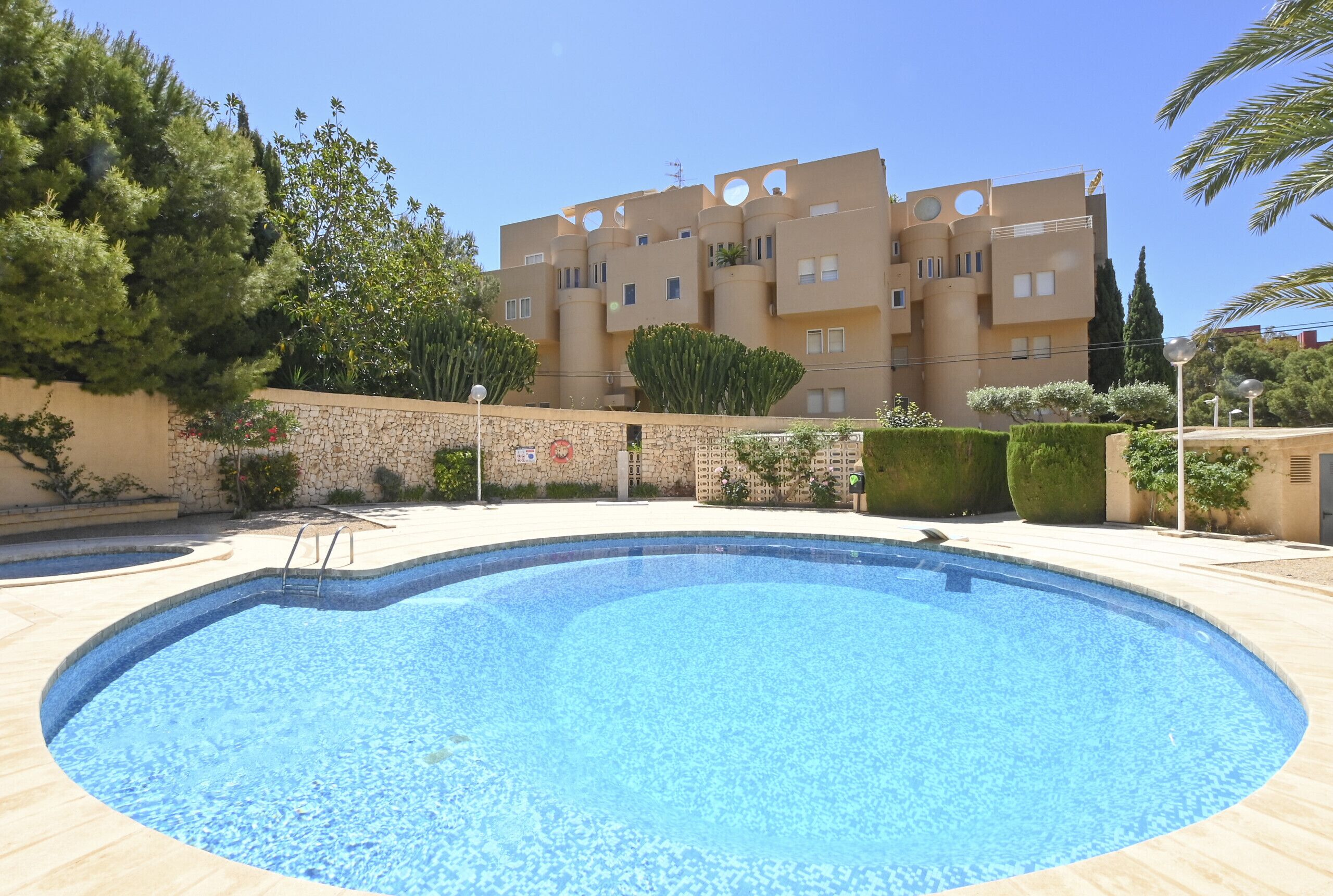 A swimming pool with a stone wall and a building with circular windows and balconies in the background.