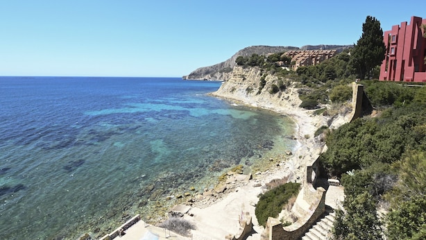 Vista costera con mar azul cristalino, costa rocosa y edificios con fachadas rojas.