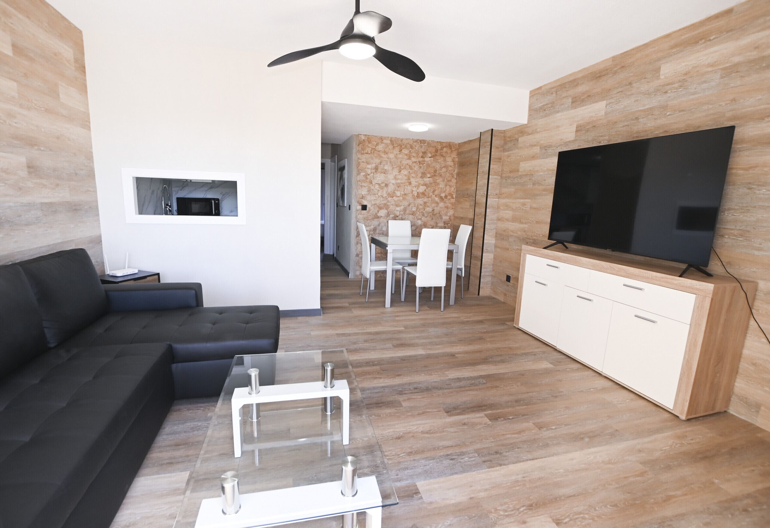 Een moderne woonkamer met een hoekbank, een glazen salontafel, een flatscreen televisie en een eethoek met witte stoelen.