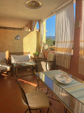 A sunlit room with a bed, a table with a plate, a chair, and a window with curtains. - Comfortable and cozy apartment in Villasimius, 5 minutes from downtown   (villasimius)