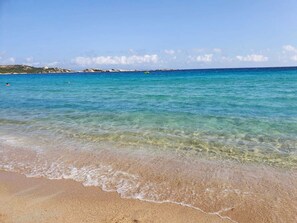 Een strand met helder turquoise water, zachte golven en veraf gelegen rotsformaties.