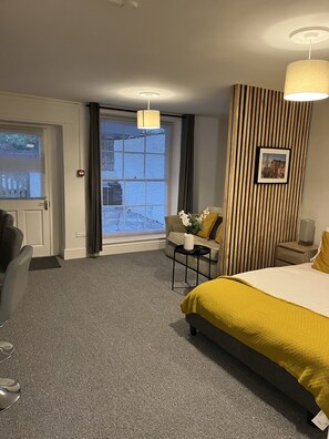 Habitación de hotel con cama, sofá, una mesita, una silla, escritorio, lámpara, un cuadro en la pared y ventana con persianas.