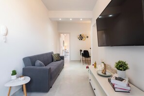 A modern living room with a grey sofa, a small white side table, a flat-screen TV, and a dining area in the background.