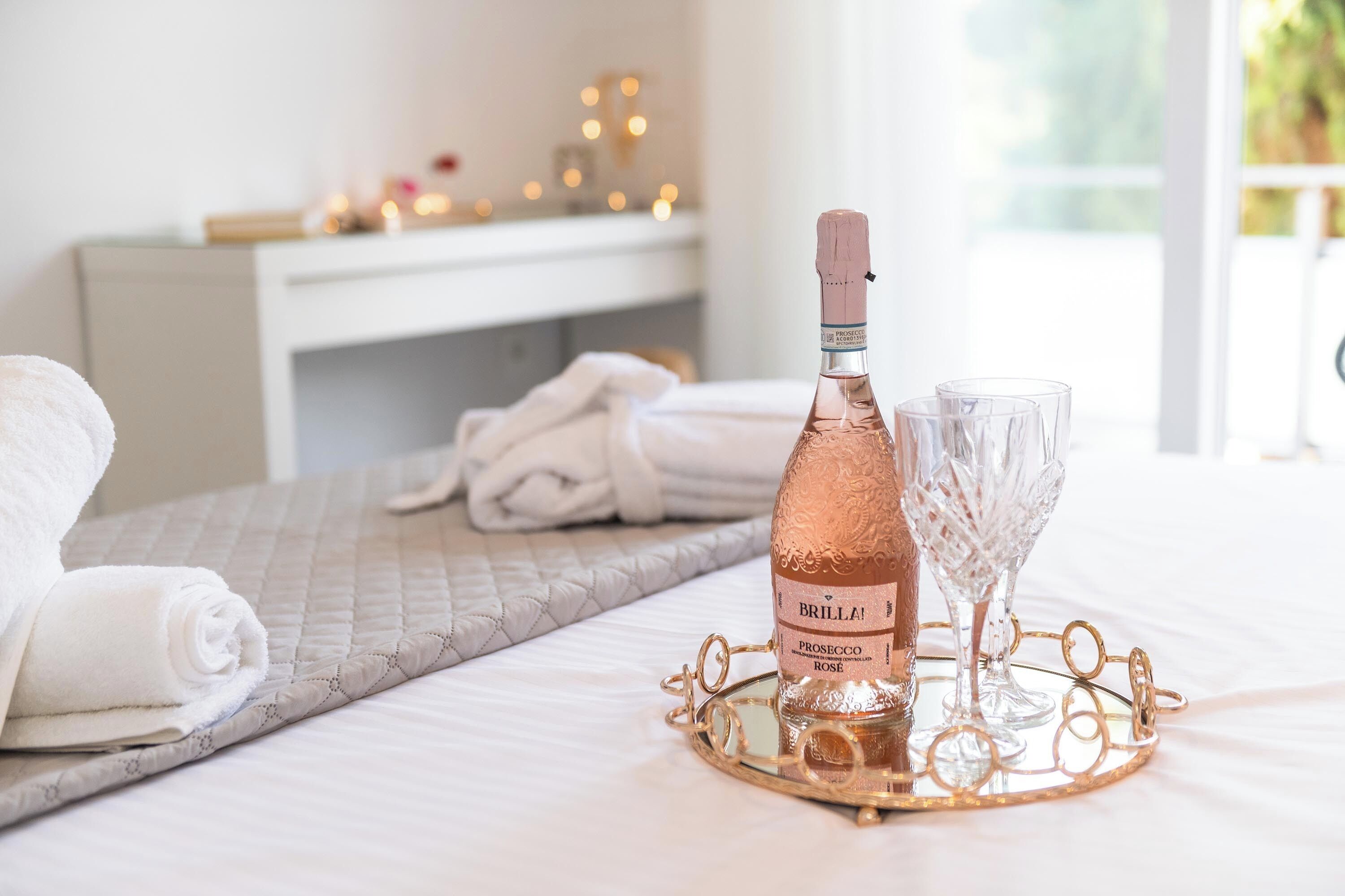 A bed with a rolled white towel, a bottle of Brilla! Prosecco Rosé, and two glasses on a tray.